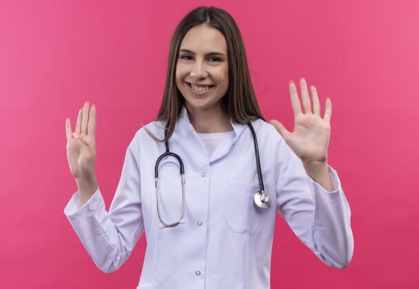礼服微笑的年轻医生女孩穿着听诊器医用长袍 在孤立的粉红色背景上显示不同的数字不同医疗年轻