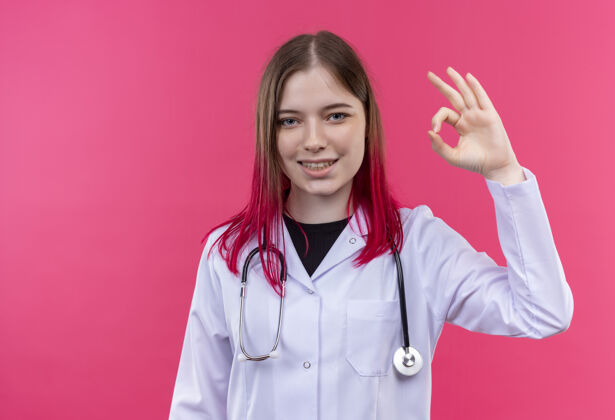 微笑微笑着的年轻医生女孩穿着听诊器医用长袍 在粉色孤立的背景上显示出良好的姿态长袍穿着年轻