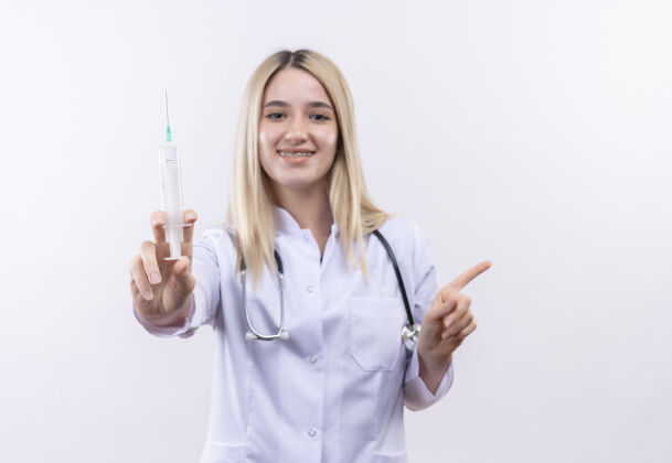 年轻微笑的医生年轻的金发女孩戴着听诊器和医用长袍戴着牙套拿着注射器指向侧面在孤立的白色背景上支架医生听诊器