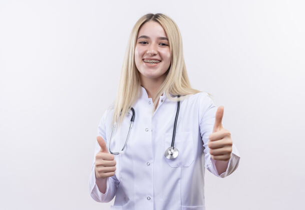 长袍微笑的医生年轻的金发女孩戴着听诊器和医用长袍 戴着牙套 在孤立的白色背景上竖起大拇指白人听诊器支架
