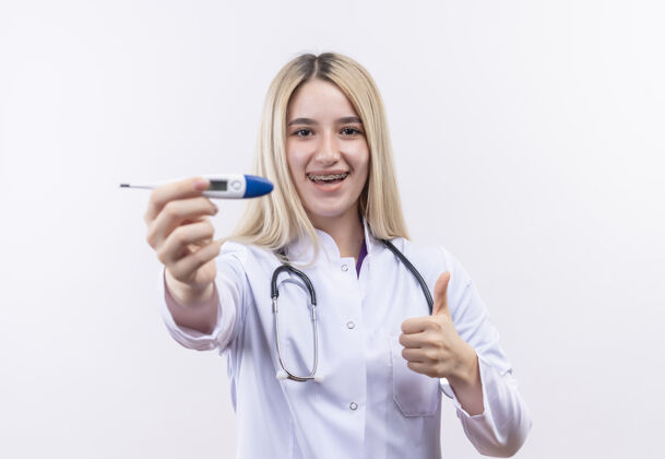 抱着微笑的医生年轻的金发女孩 戴着听诊器和医用长袍 戴着牙套 拿着温度计对着摄像机 在孤立的白色背景上竖起大拇指金发医疗听诊器