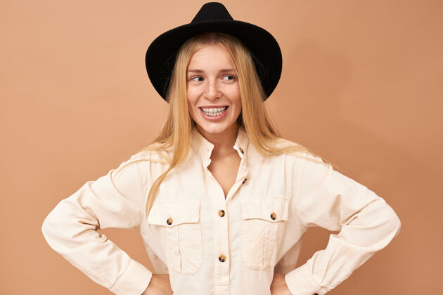 肖像可爱迷人的年轻白种女性的肖像 戴着黑色的帽子 独立地摆出自信坚定的姿势 双手放在腰上 面带微笑兴奋黑色牙齿
