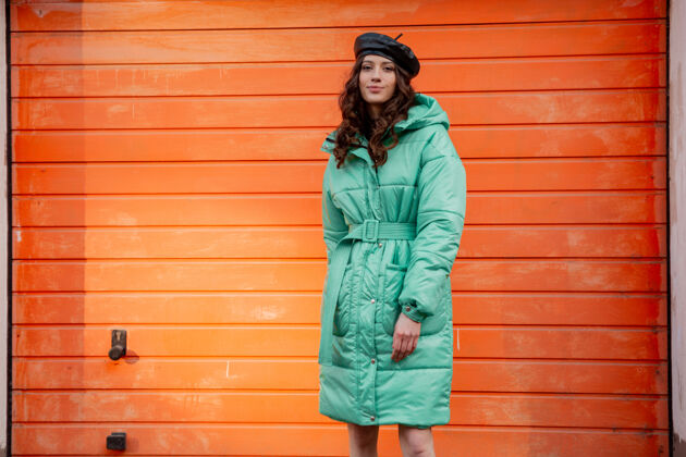 女人穿着冬秋时尚潮流蓬松外套和帽子贝雷帽的时髦女人在街上对着橙色的墙壁摆姿势风格休闲街头