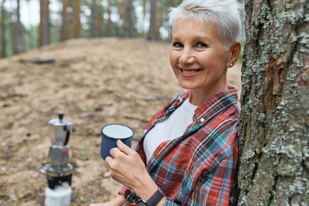 茶一幅精力充沛的退休妇女的画像 她倚靠在松树上 拿着茶杯 喝着她用野营炉上的水壶煮的茶饮料人女人