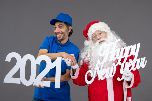 男人圣诞老人的正面图 男信使在灰色的墙上举着新年快乐和2021年的木板举行服装圣诞老人
