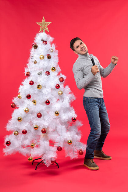 季节圣诞节的气氛与自信的家伙穿着牛仔裤站在装饰圣诞树附近 手持麦克风雪人装饰溜冰