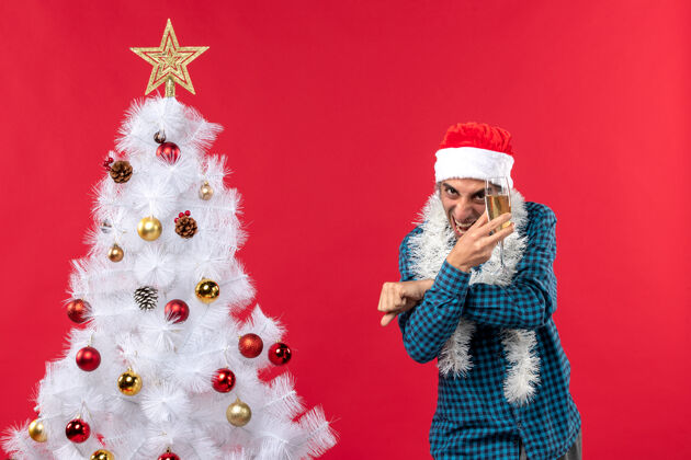 圣诞老人圣诞心情与快乐疯狂的年轻人在圣诞树附近举起一杯葡萄酒圣诞老人帽子在一个蓝色的剥夺衬衫帽子圣诞老人快乐