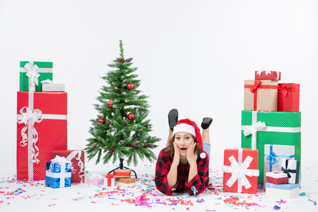 冬青年轻女子躺在白色墙壁上的圣诞礼物和小圣诞树周围的正视图礼物礼物十二月