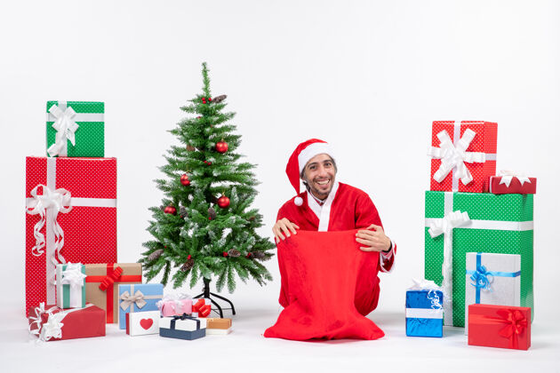 礼物笑容满面的年轻人装扮成圣诞老人 拿着礼物和装饰好的圣诞树坐在地上 在白色背景上对着镜头摆姿势装饰人地