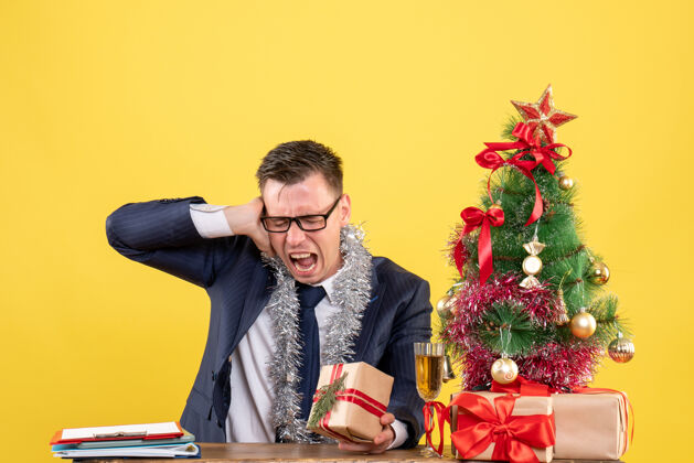 耳朵一个戴着眼镜的年轻人举着耳朵坐在圣诞树旁的桌子旁 用黄色做礼物办公室年轻人黄色