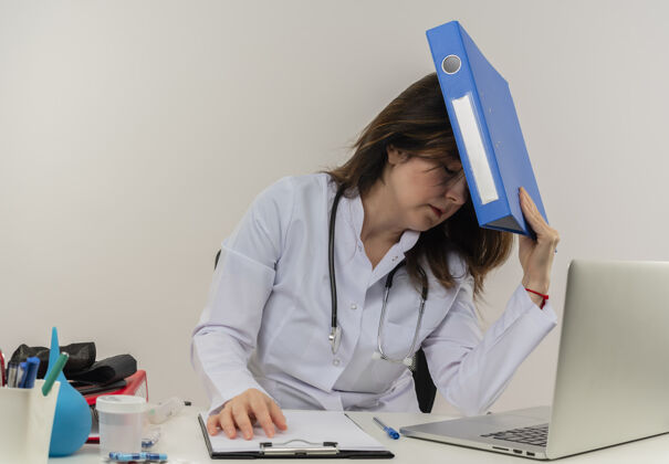 累了疲惫的中年女医生穿着医用长袍 带听诊器 坐在办公桌旁 用笔记本电脑和医疗工具 把文件夹放在头上 在隔离的白色背景上 留有复印空间坐着长袍工作