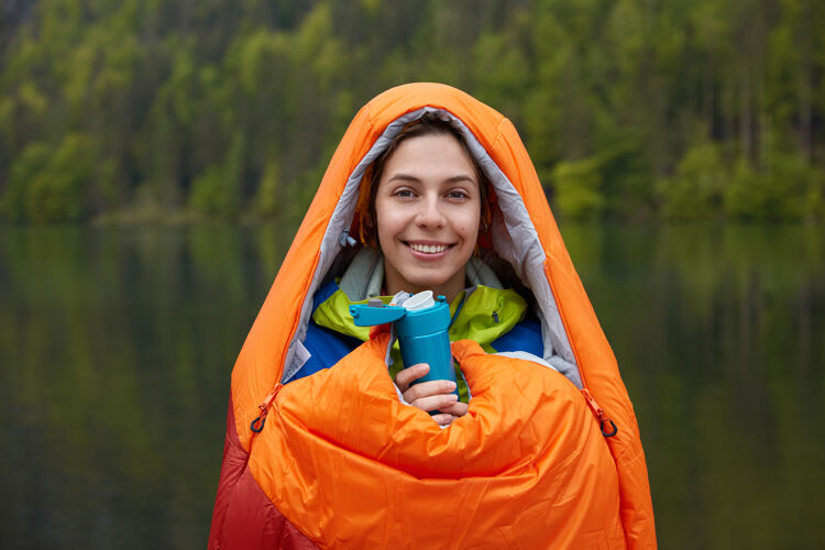 积极笑容可掬的女游客 心情舒畅 裹着睡袋 在大自然中消遣 拿着热水瓶风景休息乐观