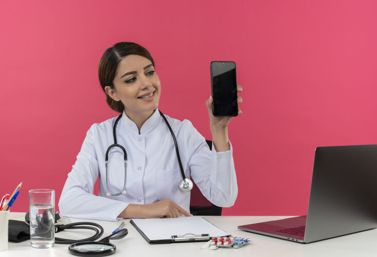 拿着微笑着年轻的女医生穿着医用长袍带听诊器坐在办公桌上用电脑拿着医疗工具拿着电话带复印空间办公桌电话听诊器