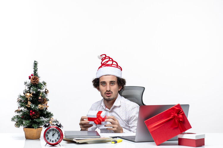 礼物新年心情与悲伤不满的年轻商人圣诞老人的帽子坐在办公室里 拿着他的礼物白色背景商人悲伤办公室