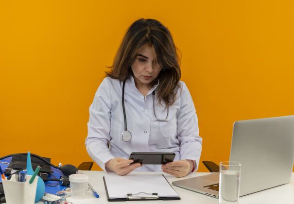 电话想一想中年女医生穿着医用长袍 带听诊器 坐在办公桌上 拿着笔记本电脑 带着医疗工具 拿着手机 看着孤立的橙色背景 还有复印空间拿着穿着长袍