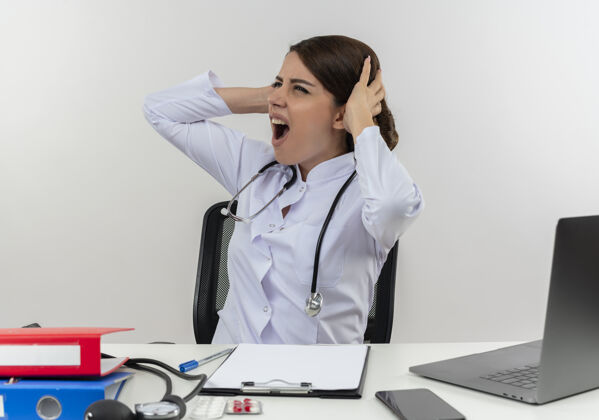 工作压力重重的年轻女医生穿着医用长袍 手持听诊器坐在办公桌旁 用医疗工具在电脑上工作 用复印空间抓住头部穿着听诊器压力