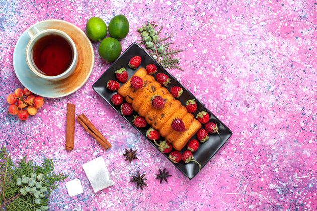 顶部顶视图美味的烤蛋糕放在黑色蛋糕锅里 粉红色的桌子上放着新鲜的红色草莓茶和柠檬新鲜水果烘焙