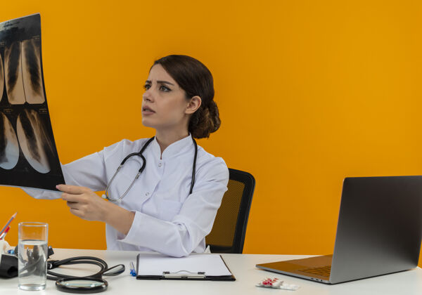 工作想着年轻的女医生穿着医用长袍 带着听诊器坐在办公桌旁 拿着医疗工具 拿着电脑 在隔离的黄色背景上看x光片听诊器电脑办公桌