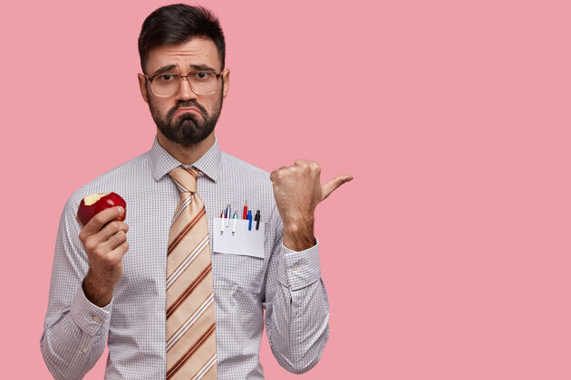 苹果不满的上班族吃苹果 皱眉头 用拇指指着旁边 穿着正式的衬衫和领带皱眉男沮丧