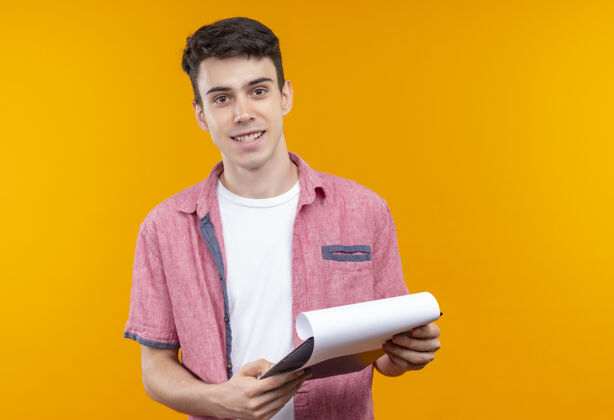 橙色笑容满面的白人年轻人穿着粉色衬衫拿着剪贴板在孤立的橙色背景上微笑穿着衬衫
