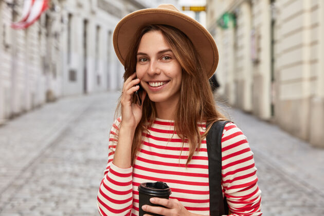 帽子开朗的女游客在城市空间摆姿势 用纸杯喝外卖咖啡女性手机交谈