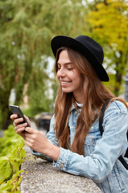 手持开朗时尚女人垂直镜头读搞笑短信 用手机应用 在户外绿树上摆姿势无线应用程序牛仔