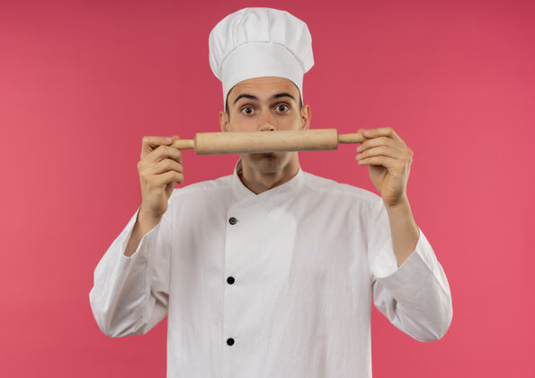 制服惊讶的年轻男厨师穿着厨师制服拿着擀面杖围在脸上围着拿着厨师
