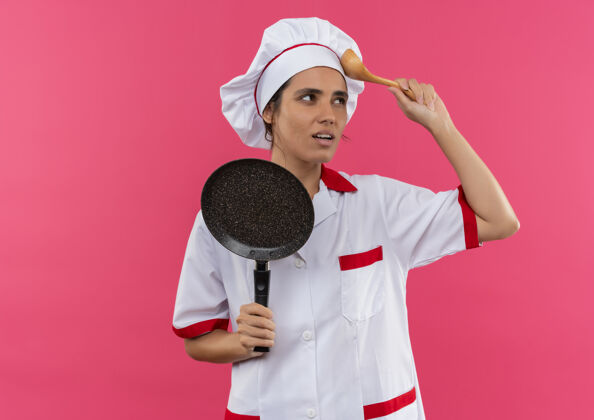 穿想年轻的女厨师穿着厨师制服拿着煎锅把勺子放在额头上和复印空间拿煎思考