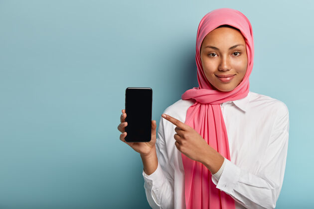 头巾买这个设备高兴黑皮肤的女人在粉红色面纱 在智能手机点促销自由空间指示