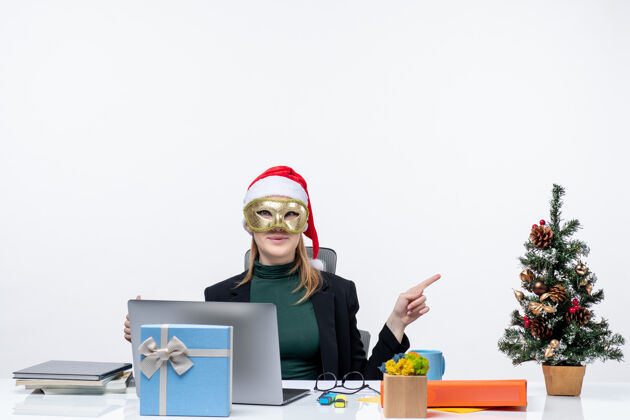 工作圣诞气氛 戴着圣诞老人帽 戴着面具的年轻女子坐在桌子旁 指着白色背景上的东西人电脑办公室