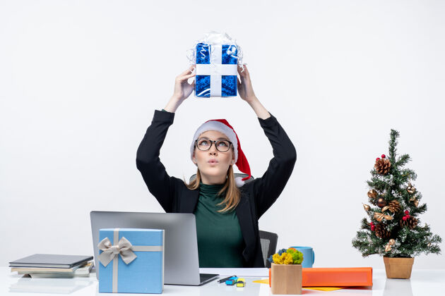 年轻人带着圣诞老人帽子戴着眼镜的年轻女子坐在桌子旁 头上戴着白色的背景 脸上挂着礼物办公室帽子坐着