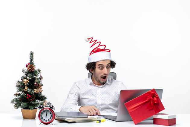 微笑快乐的积极震惊年轻的商人与有趣的圣诞老人帽子仔细检查他在白色背景的办公室商人帽子办公室