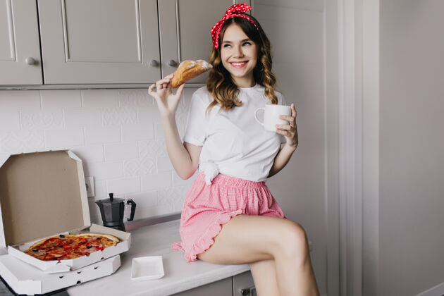 微笑穿着粉色短裤的快乐女孩吃羊角面包留着卷发的幽默女模特喝着披萨茶晚餐披萨年轻