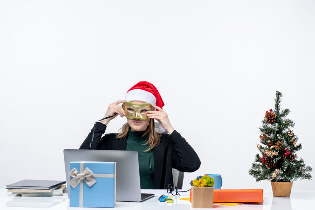 电脑一个戴着圣诞老人帽子戴着面具的积极的年轻女人坐在一张桌子旁 桌子上有一棵圣诞树和一份白色背景的礼物面具帽子圣诞老人