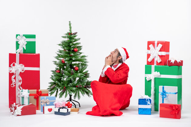 礼物被吓得目瞪口呆的年轻人打扮成圣诞老人 拿着礼物和装饰好的圣诞树坐在白色背景的地上礼物年轻人礼物