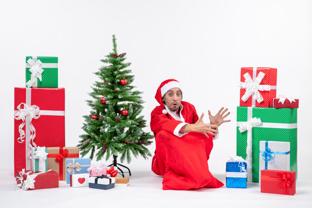 人惊讶的年轻人打扮成圣诞老人和礼物装饰圣诞树坐在地上的白色背景礼物坐着装饰