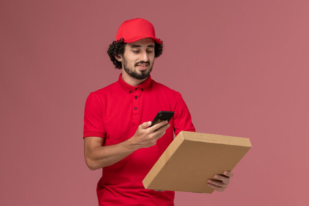 空的正面图穿着红色衬衫和斗篷的男信使拿着空的送货食品盒在粉红色的墙上拍照服务拿着视图