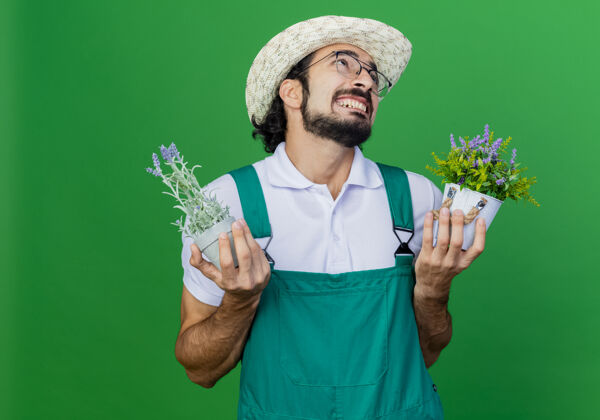 抱着年轻的留着胡须的园丁 穿着连体衣 戴着帽子 手里拿着盆栽植物 带着恼怒的表情抬起头来绿色年轻植物