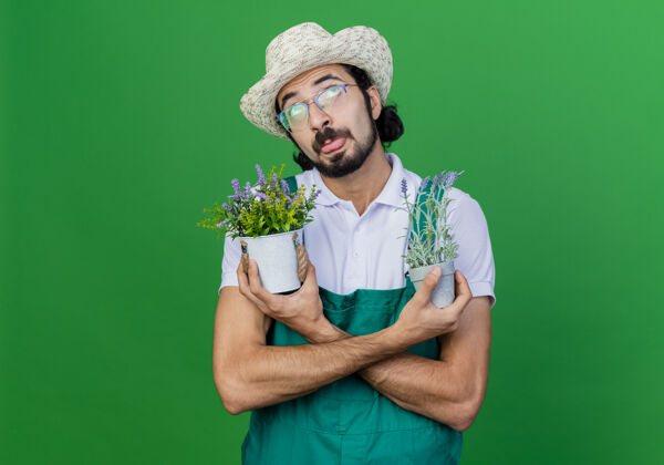 植物有趣的年轻留着胡须的园丁 穿着连体衣 戴着帽子 手里拿着盆栽植物 抬起头 伸出舌头园丁向上绿色
