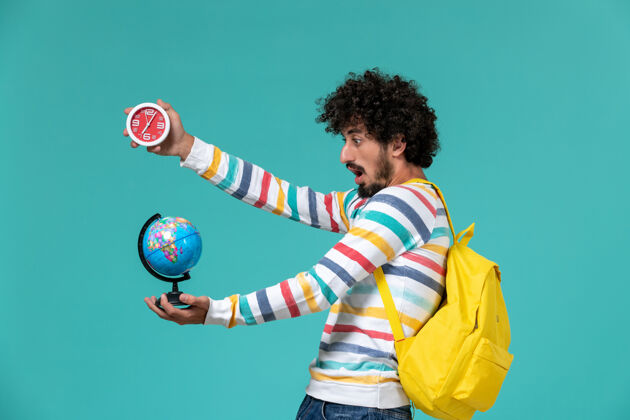 男性穿着条纹衬衫 背着黄色背包 蓝色墙上拿着小地球仪和钟的男学生的正面图地球舞会大学