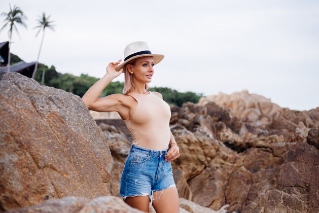 户外穿着米色紧身衣蓝色牛仔短裤和经典时尚帽子的欧洲年轻女子的户外时尚写真在热带岩石海滩温暖的夕阳下女人女士肖像