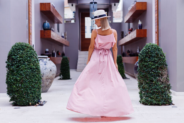 优雅穿着浪漫优雅粉色长裙的白种女人的肖像 在豪华的别墅酒店度假 美丽的热带棕榈树映衬着戴着经典白帽子的女人配件度假露台