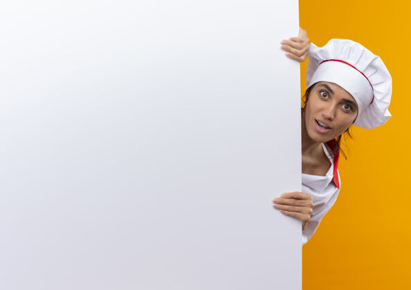 穿着惊讶的年轻女厨师穿着厨师制服拿着白色的墙壁与复制空间墙惊讶厨师