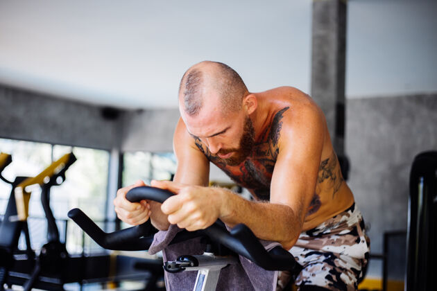 训练纹身 肌肉发达 胡须浓密的男人骑着自行车在体育馆里锻炼有氧运动 靠近大窗户 外面可以看到树运动健康俱乐部骑马