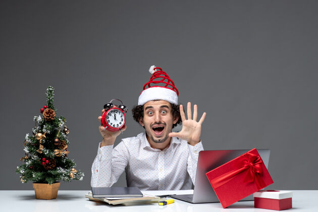 商务年轻人戴着圣诞老人的帽子 拿着钟 在黑暗的背景下 在办公室里展示五个 这让生意人大吃一惊圣诞老人帽子办公室