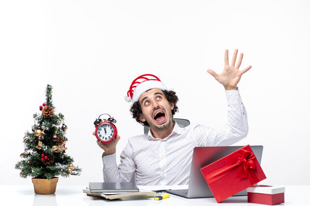 微笑年轻愤怒的商人戴着圣诞老人的帽子 拿着时钟 在白色背景下展示五个坐在办公室里的人商务人士人年轻