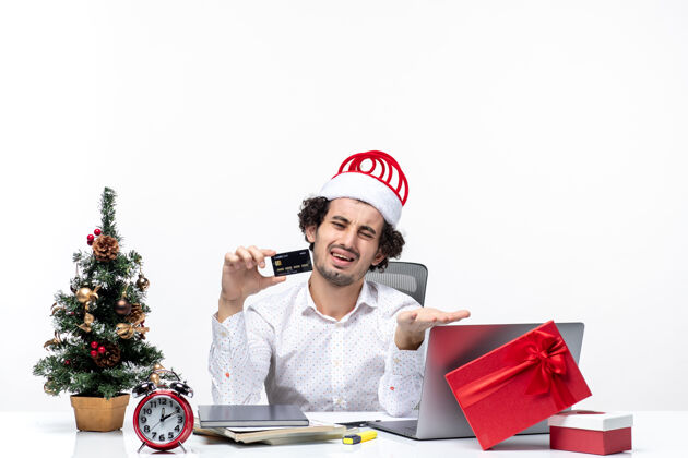 圣诞老人节日喜庆心情带着年轻疲惫愤怒的商务人士戴着圣诞老人的帽子 拿着他的银行卡在白色背景的办公室里拍照留念股票商务年轻