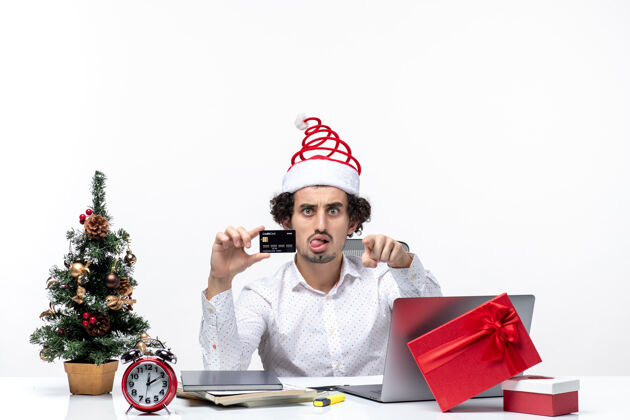 圣诞老人带着圣诞老人帽子 拿着银行卡 在白色背景上指着办公室里的东西的年轻商人的节日喜庆心情银行圣诞节帽子