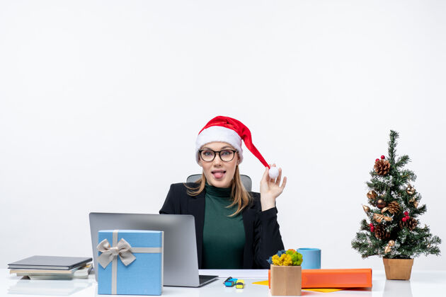 帽子一个积极的女商人坐在一张桌子旁 手里拿着圣诞树和礼物 手里拿着圣诞老人的帽子 在白色的背景上伸出舌头女士桌子圣诞