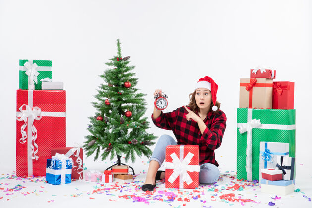 礼物前视图的年轻女子坐在圣诞礼物周围拿着白色墙上的时钟装饰女人圣诞节